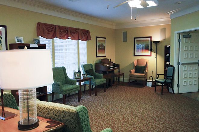A cozy living room area with green chairs and a sofa arranged around a carpeted floor, featuring ambient lighting and windows with curtains.
