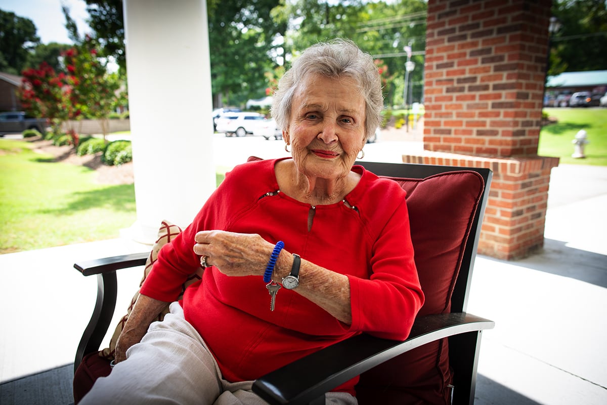 An elderly woman in a red shirt is sitting on a porch, smiling at the camera.