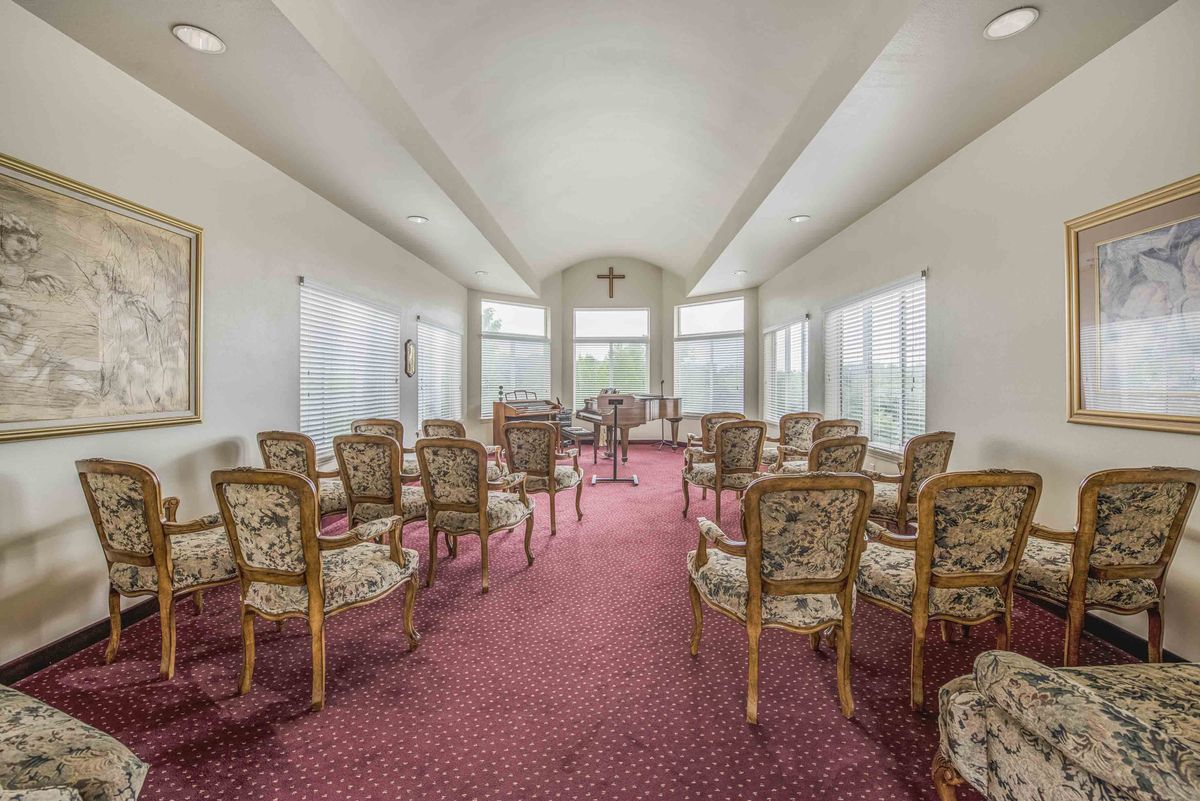 A well-lit room with ornate chairs arranged in rows on a red carpet, featuring large windows and high ceilings.