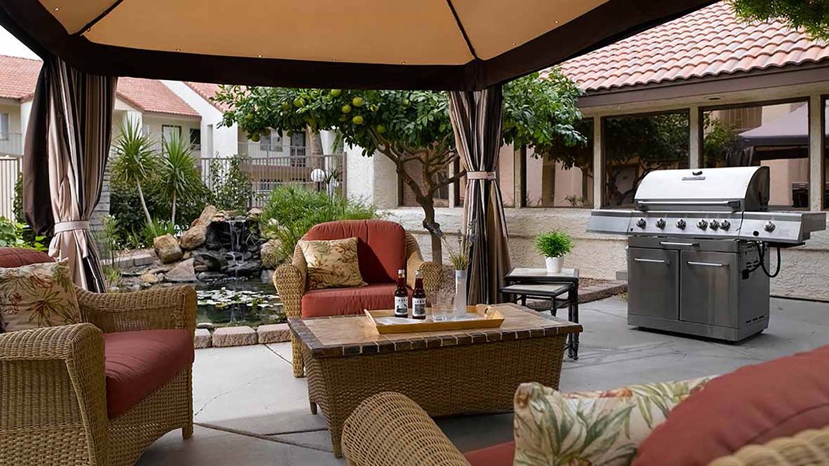 A cozy outdoor patio with wicker furniture and cushions under a gazebo, surrounded by a garden setting.