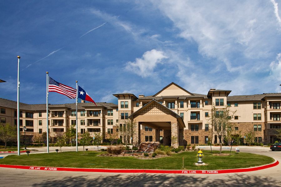 A modern building with American and Texas flags displayed at the entrance, surrounded by well-maintained landscaping under a clear blue sky.