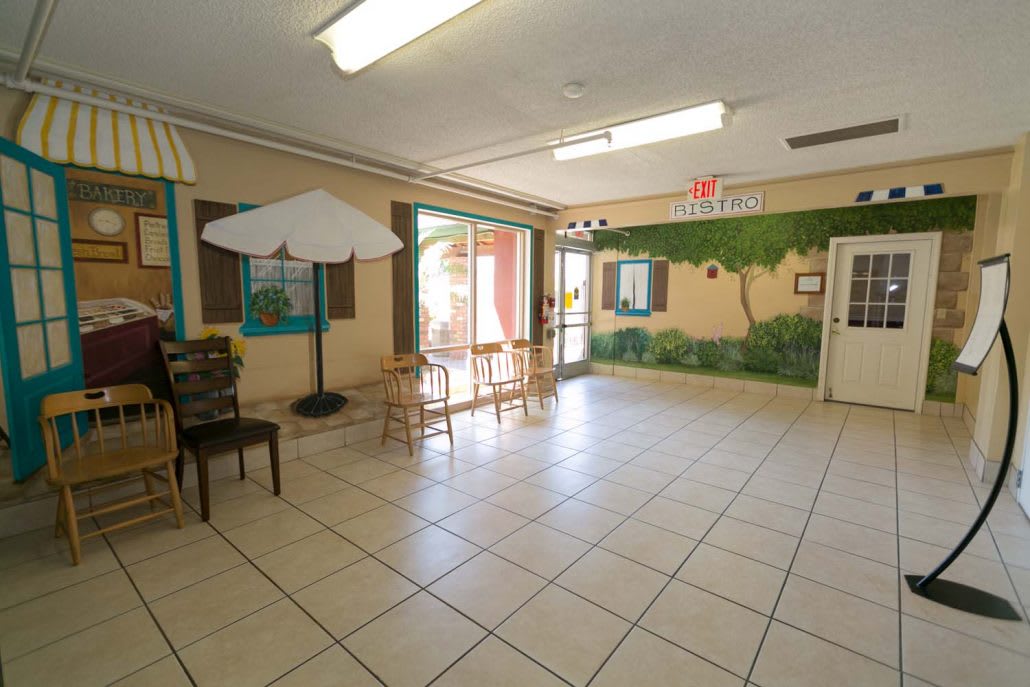 A bright waiting area with tiled flooring, tables, chairs, an umbrella, and a mural on the wall.
