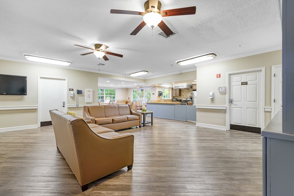 A spacious living room with wooden flooring, featuring sofas and ceiling fans, illuminated by natural light through large windows.