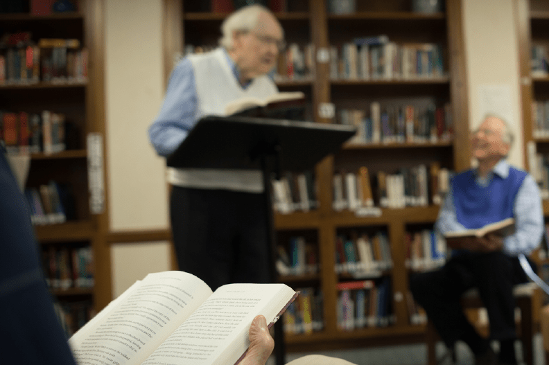 A man standing at a podium reads to an audience in a library setting.