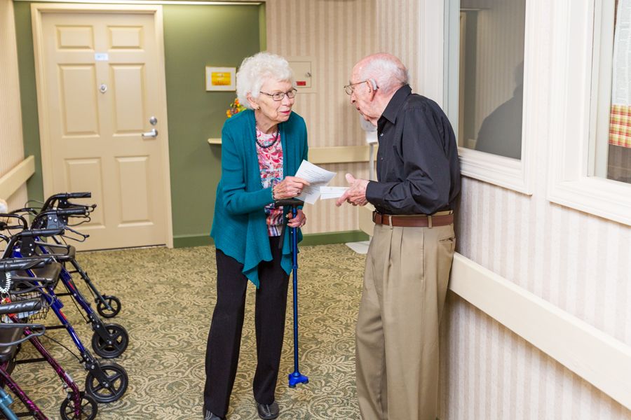 Two elderly individuals engage in a conversation indoors, one using a walking stick.