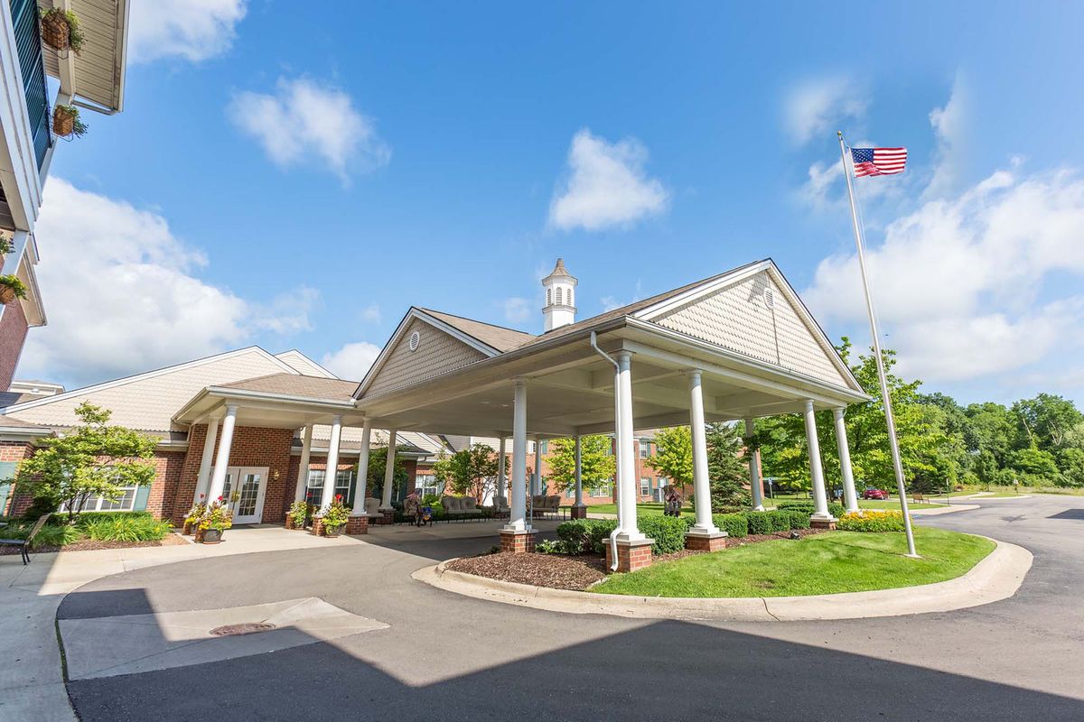 The entrance of a building with a covered driveway and columns, featuring an American flag against a blue sky with clouds.