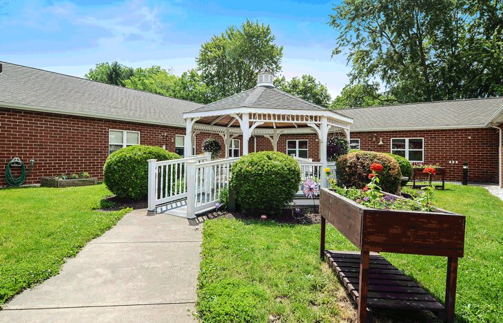 A charming gazebo in a garden setting with a pathway leading to it, surrounded by greenery and adjacent to a brick building.