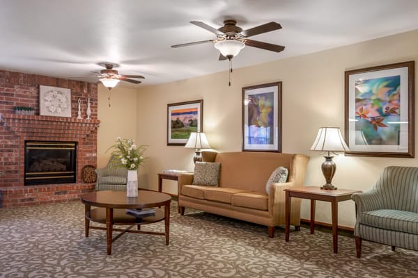 A cozy living room featuring a sofa, chairs, wooden tables, brick fireplace, ceiling fans, and warm lighting.