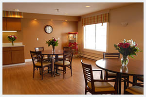 A cozy dining area with tables and chairs, a wall clock, and natural light coming through the window.