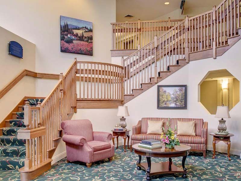 A cozy living room with a staircase, featuring a pink armchair and sofa, a round coffee table, and decorative paintings on the walls.