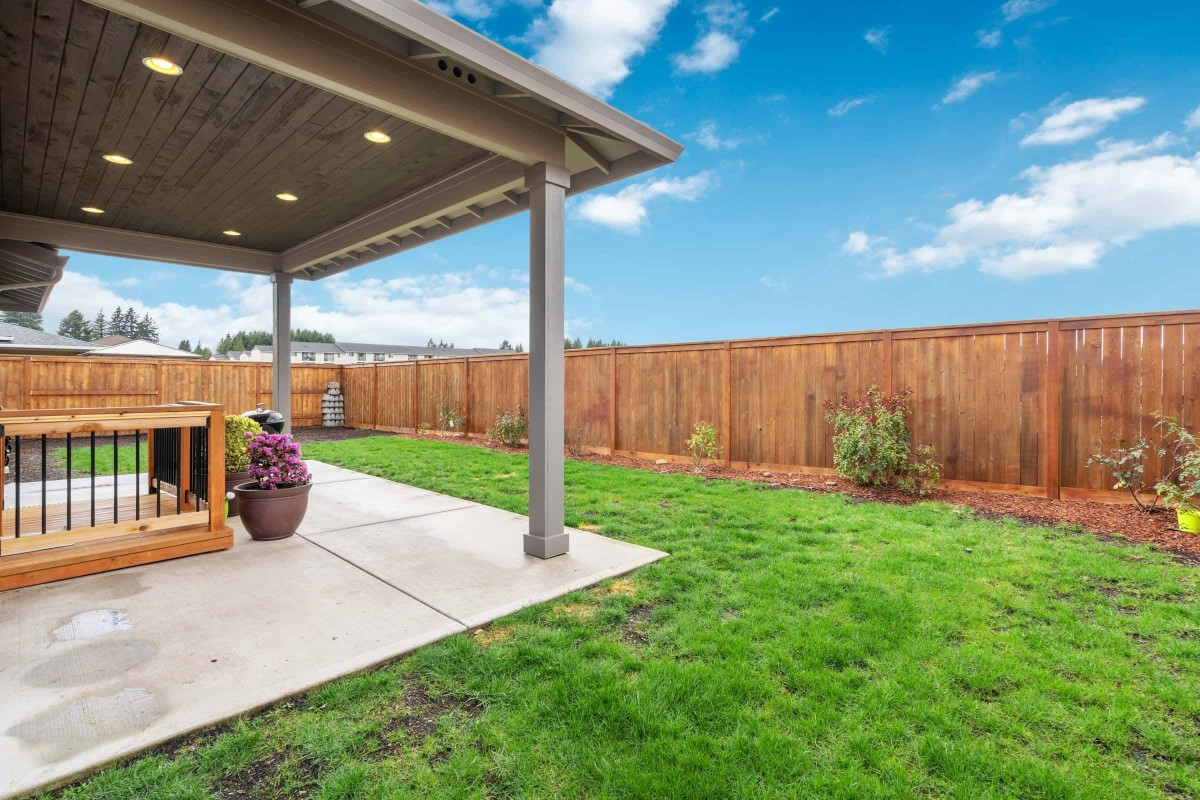 A backyard with a covered patio, grassy lawn, and wooden fence under a clear blue sky.