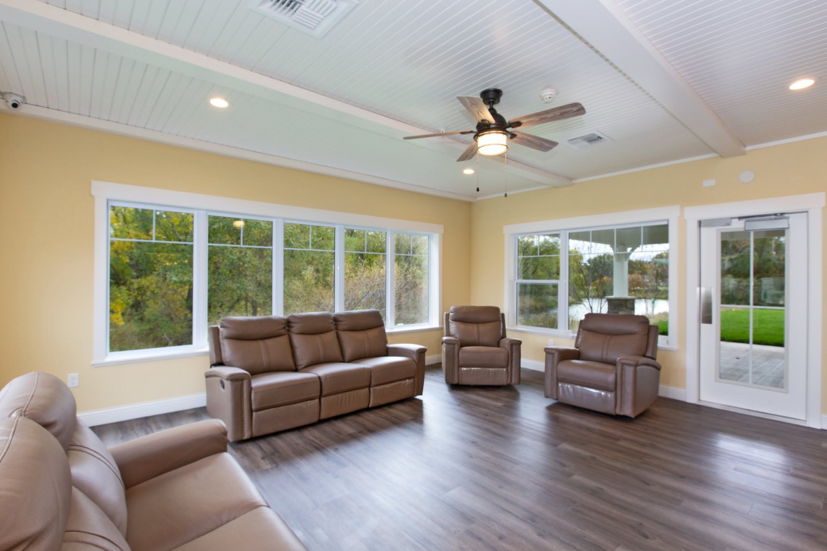 A spacious living room with large windows, brown leather furniture, and a ceiling fan.