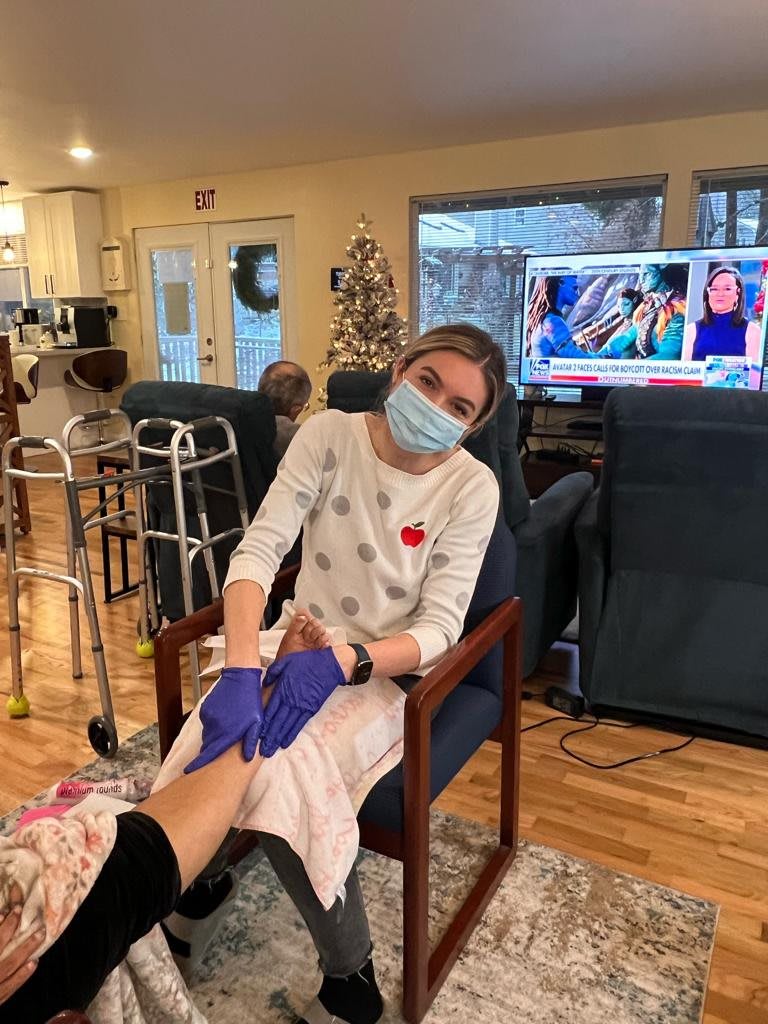 A caregiver wearing a mask and gloves tends to a patient in a nursing home setting, with a television on in the background.