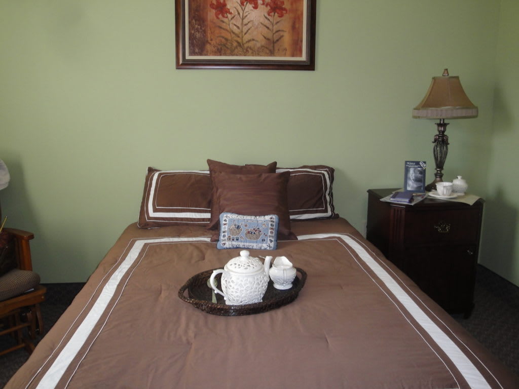 A cozy bedroom featuring a neatly made bed with a brown and white bedspread, adorned with a tray holding a tea set. A framed artwork hangs on the light green wall above, and a bedside table with a lamp adds to the room's decor.