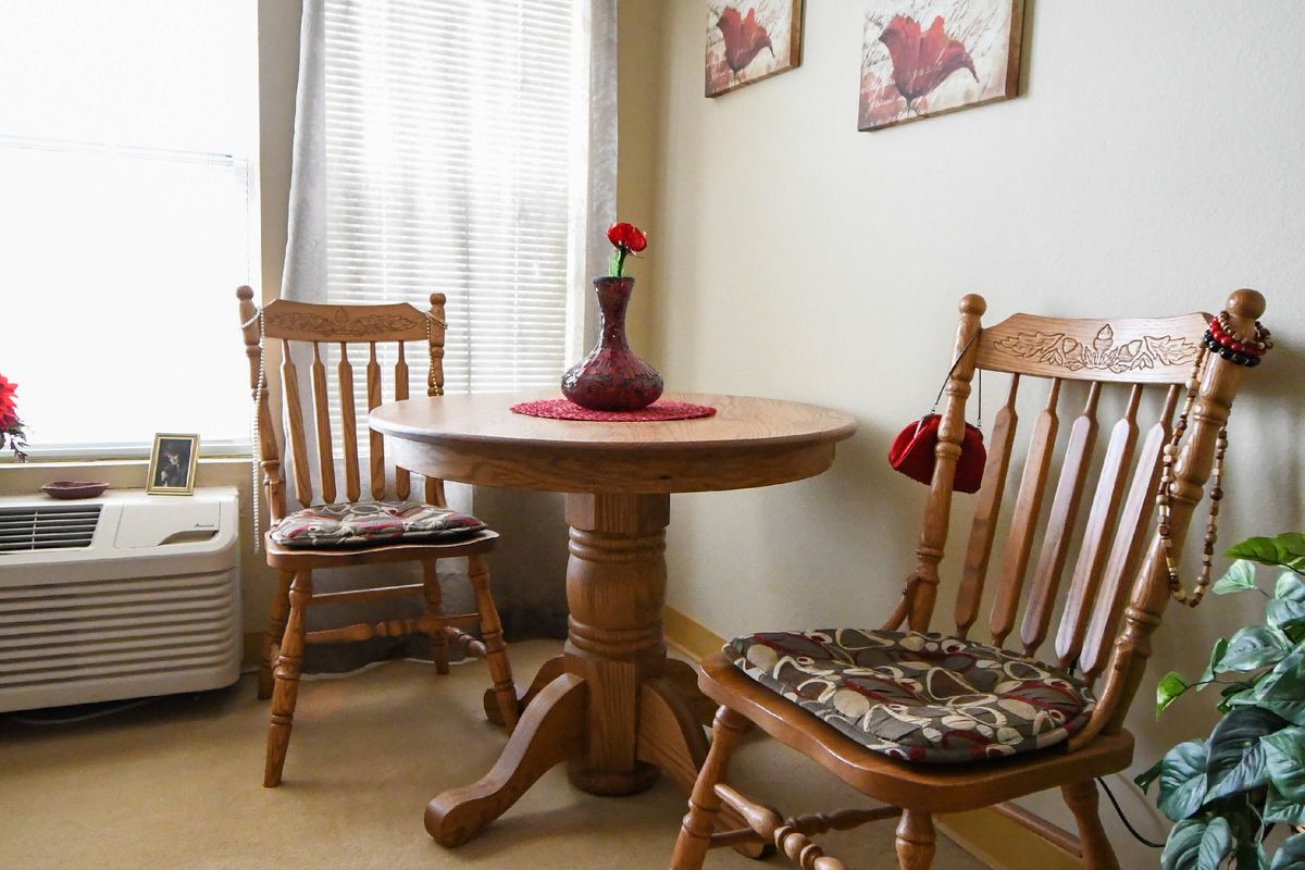 A cozy dining nook featuring a wooden table with two matching chairs, adorned with cushions and a red vase holding a single flower.