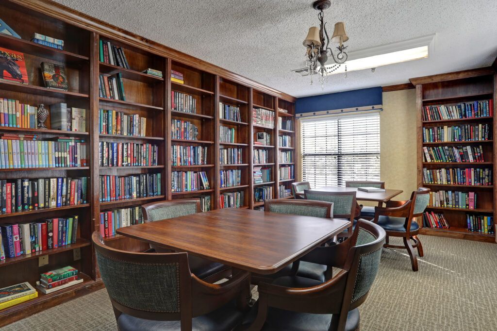 A cozy library room with bookshelves, a central wooden table surrounded by chairs, and natural light coming through the window.