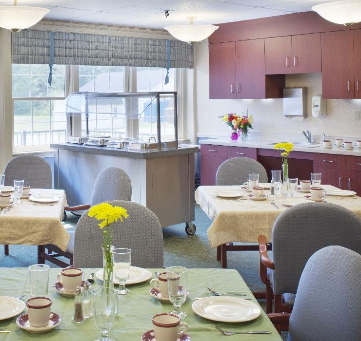 A cozy dining room with neatly set tables, complete with floral decorations and a kitchen area in the background.