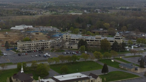 An aerial view of an urban area featuring multiple buildings, a parking lot, and patches of green space under an overcast sky.