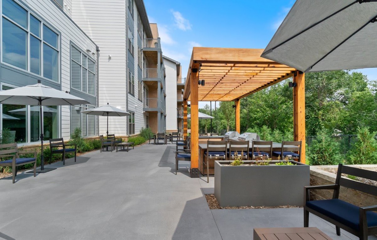 A modern residential outdoor patio with a wooden pergola, featuring tables and chairs under a clear blue sky.