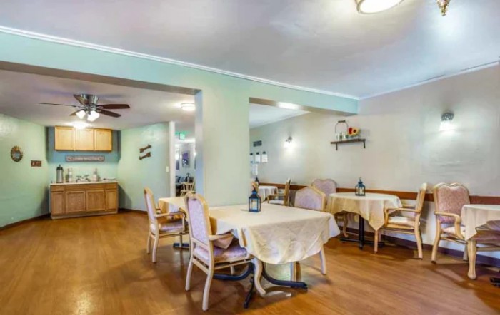 A cozy dining area with neatly arranged tables and chairs, decorated with tablecloths and decorative bottles, featuring a warm wooden floor.