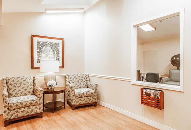 A cozy waiting room with patterned chairs and a small table, featuring a flower vase and a magazine rack under a framed painting.