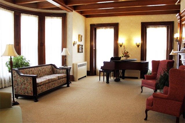 A classic-style living room featuring a sofa, armchairs, and a piano with large windows allowing natural light.