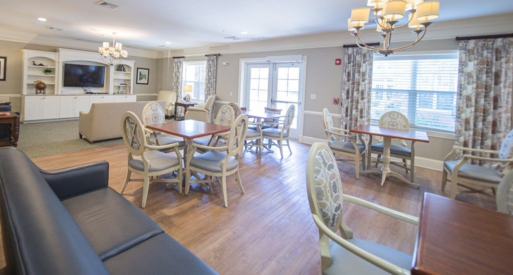 A bright and inviting dining area with multiple tables and chairs, featuring large windows and elegant chandeliers.