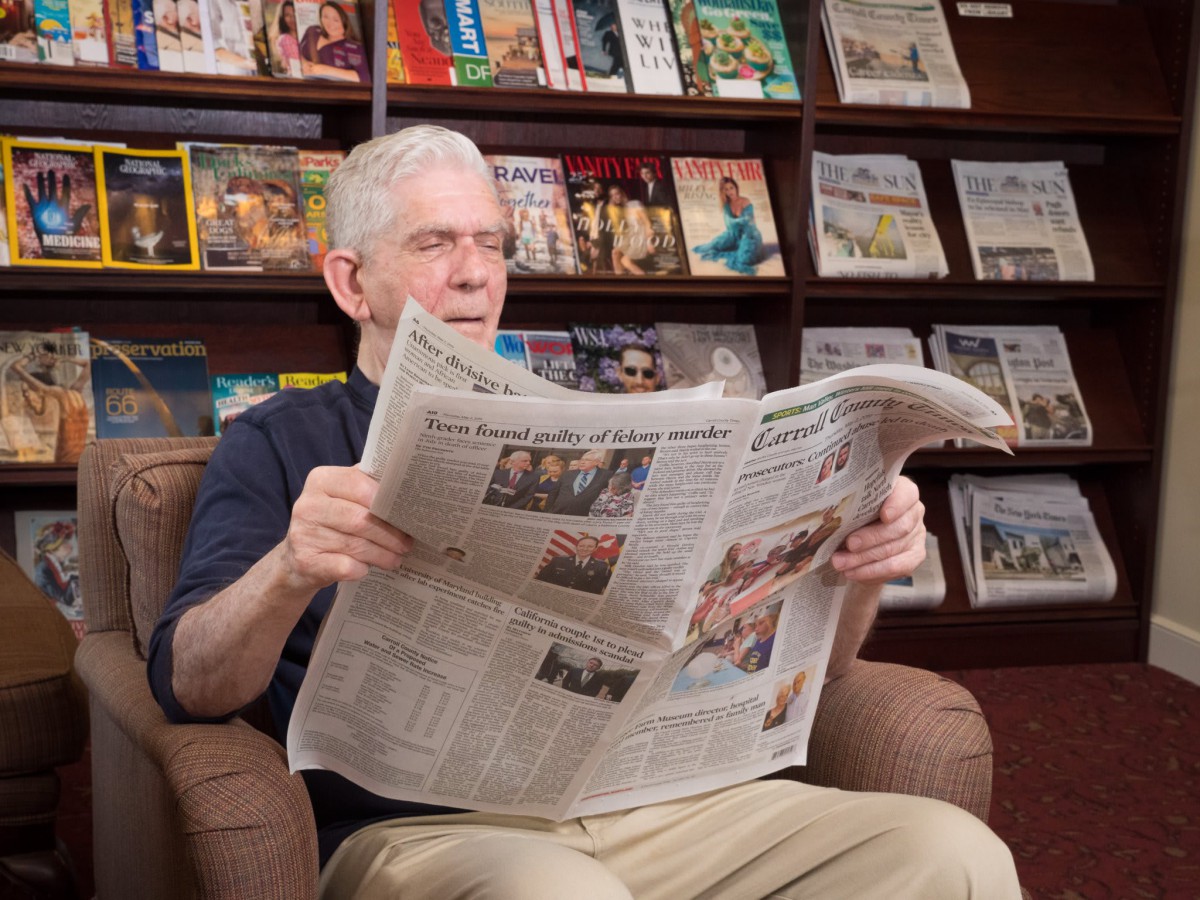 An elderly man reading a newspaper in a library surrounded by magazines.