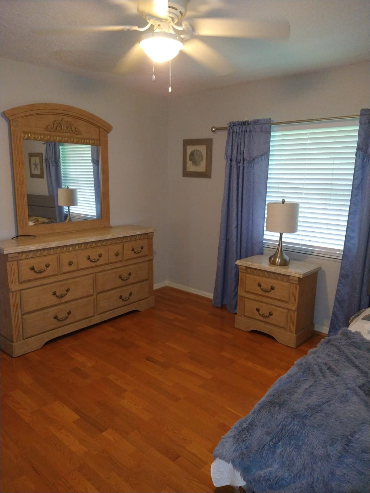 A cozy bedroom featuring a wooden dresser with a mirror, a bedside table with a lamp, and blue curtains framing the window.