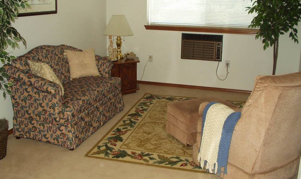 A cozy living room with a patterned sofa, an ottoman, and a floor lamp beside a window featuring an air conditioner.