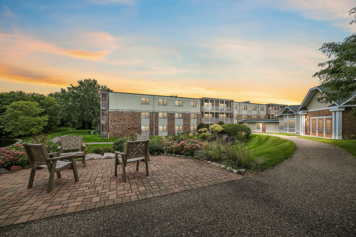 A serene outdoor view at sunset featuring a building, two chairs on a pathway, and lush greenery.