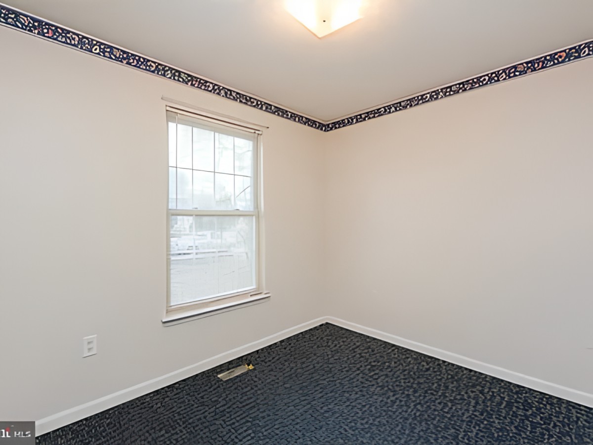 An empty room with carpet flooring and a single window, featuring a light fixture on the ceiling and decorative wall trim.