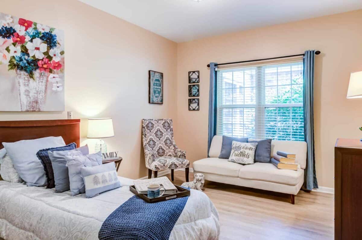 A cozy bedroom with a bed, armchair, and sofa, featuring light colors and elegant decor.