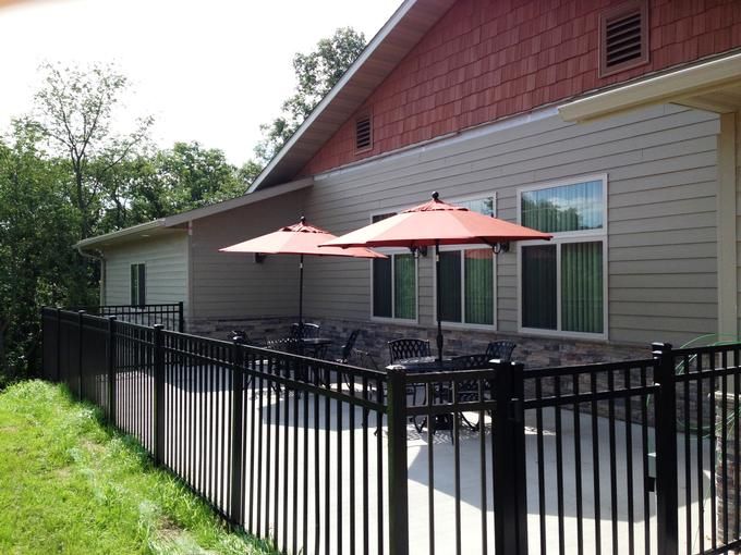 A sunny outdoor patio with tables and red umbrellas next to a building, surrounded by a black metal fence.