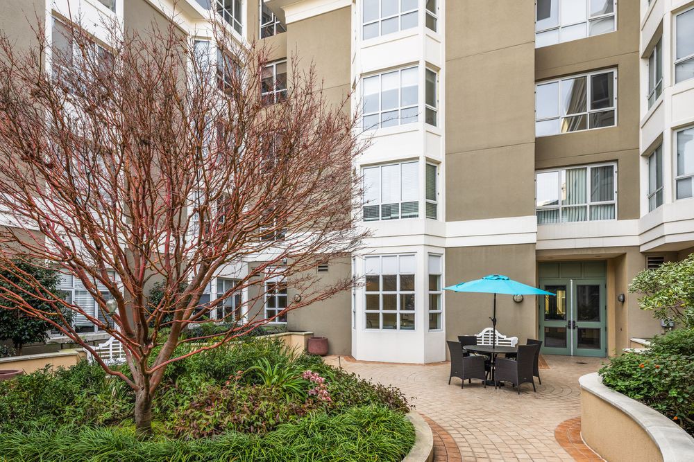 A cozy apartment courtyard with seating and an umbrella, surrounded by landscaped greenery.