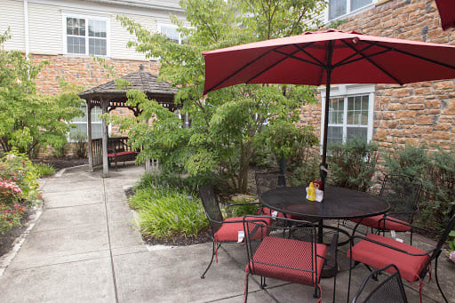 A cozy patio area with red-cushioned chairs, a matching umbrella, and a gazebo surrounded by greenery.
