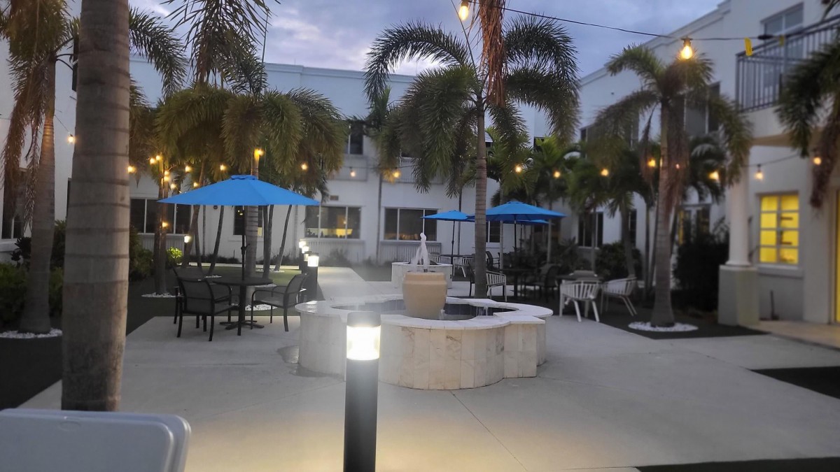 A tranquil patio setting with a fountain, palm trees, and blue umbrellas illuminated by evening lights.