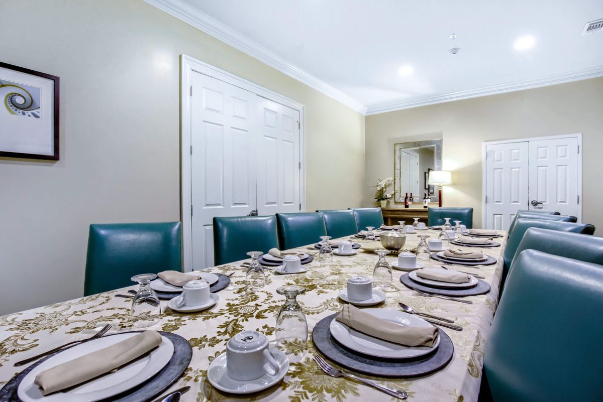 An elegantly set dining table with an ornate tablecloth, fine china, and upholstered chairs in a well-decorated room.