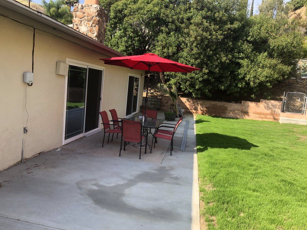 A cozy patio setup with a table and chairs under a red umbrella, situated next to a lush green lawn.