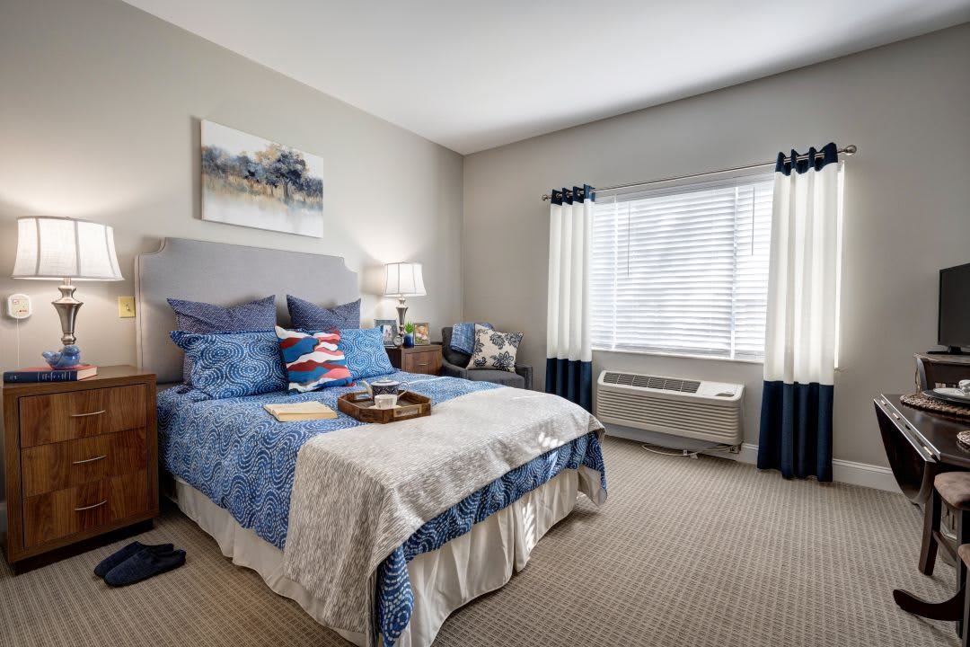 A cozy bedroom with a blue and white theme, featuring a neatly made bed, window with curtains, and an air conditioning unit.
