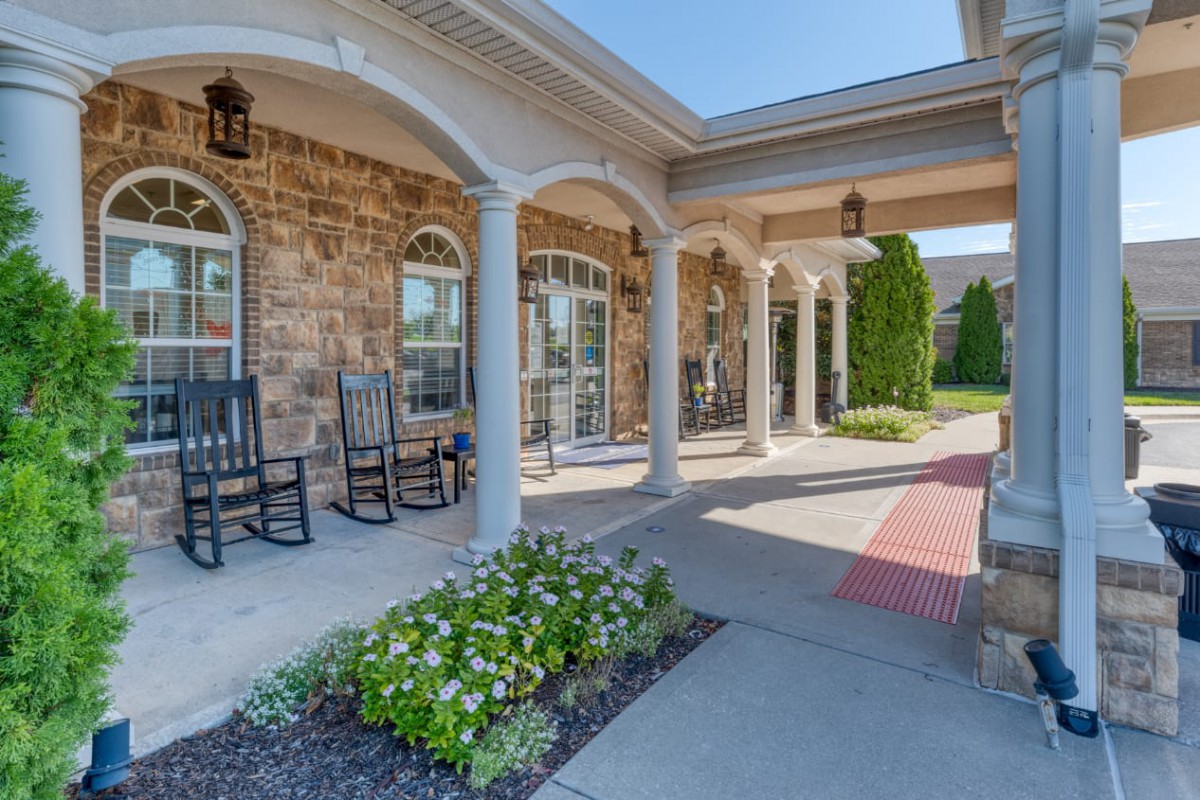 A charming porch with rocking chairs and columns, featuring a flower bed and walkway beside a brick building.