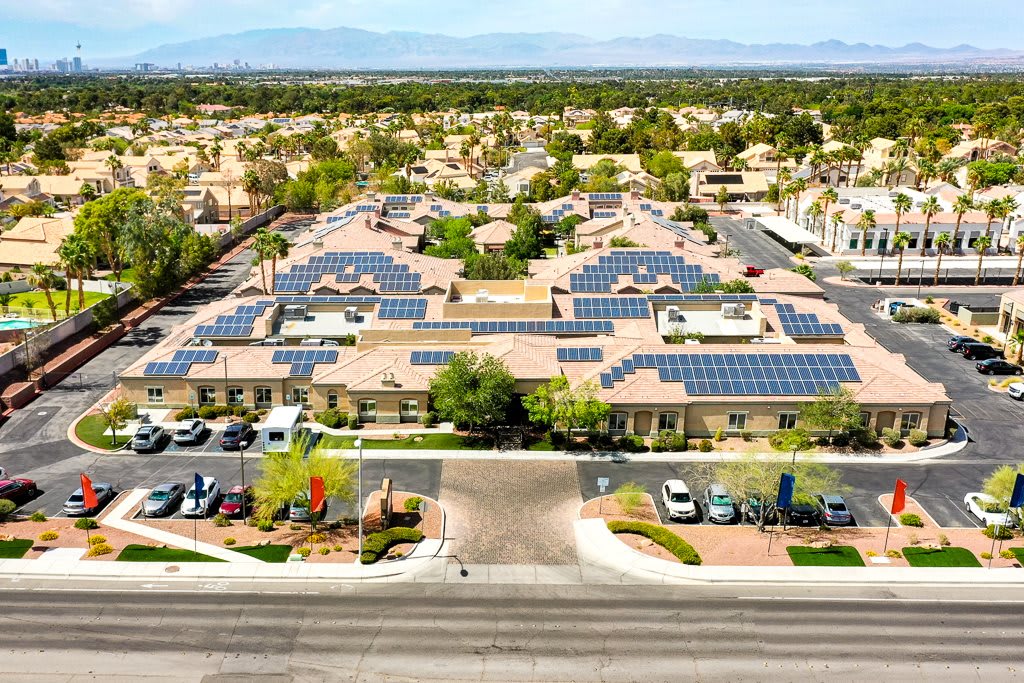 An aerial view of a complex with solar panels on the rooftops, surrounded by a parking lot and trees in a residential area.