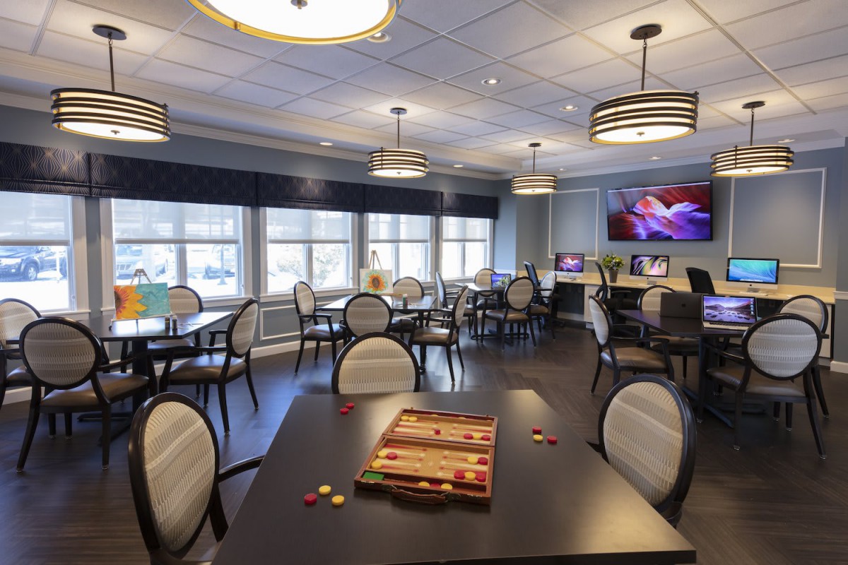 A community room with tables and chairs, featuring a backgammon board on one table, large windows, and a television on the wall.
