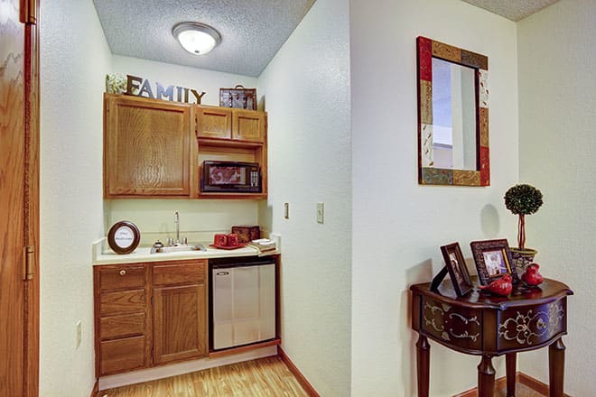 A small kitchen area with wooden cabinets and a countertop, featuring decorative items and a mirror on the adjacent wall.