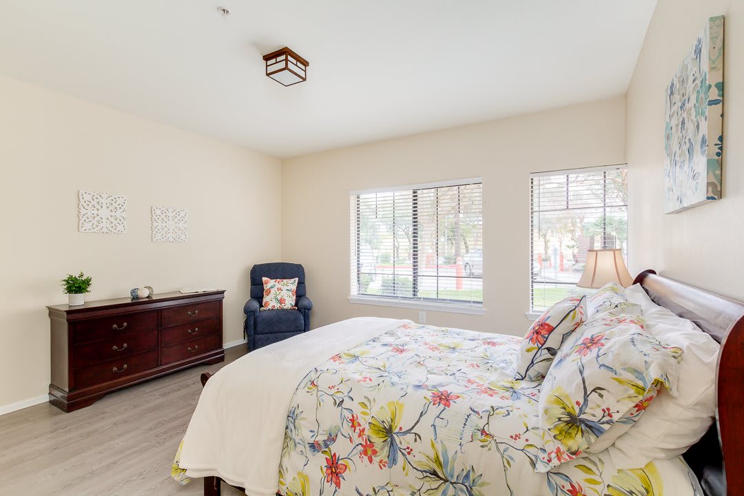 A bright and cozy bedroom with floral bedding, a dresser, and large windows.