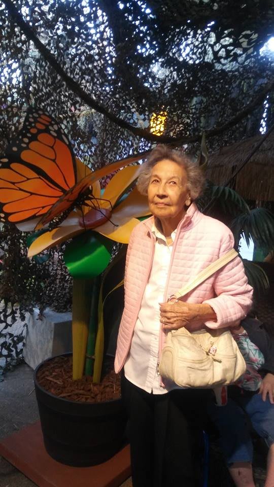 An elderly woman stands in front of a large butterfly artwork indoors.
