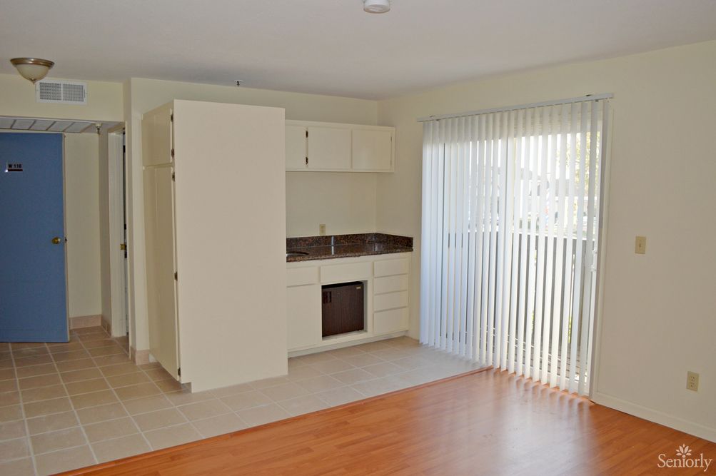 A well-lit studio apartment featuring a small kitchenette and large windows with vertical blinds.