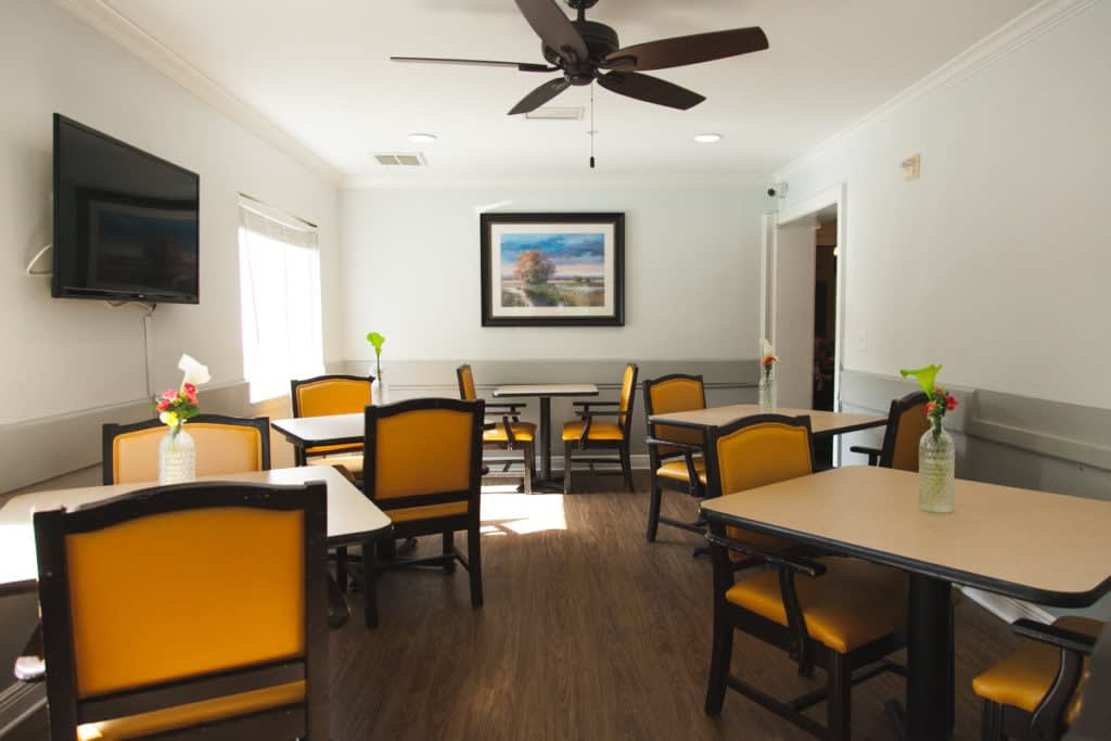A bright and inviting dining room with multiple tables and chairs, featuring a ceiling fan and a landscape painting on the wall.