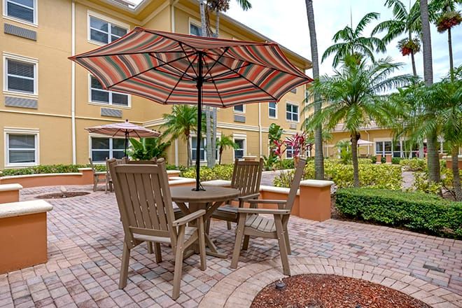 A cozy outdoor patio area with table and chairs under a striped umbrella, surrounded by lush greenery and palm trees near a residential building.