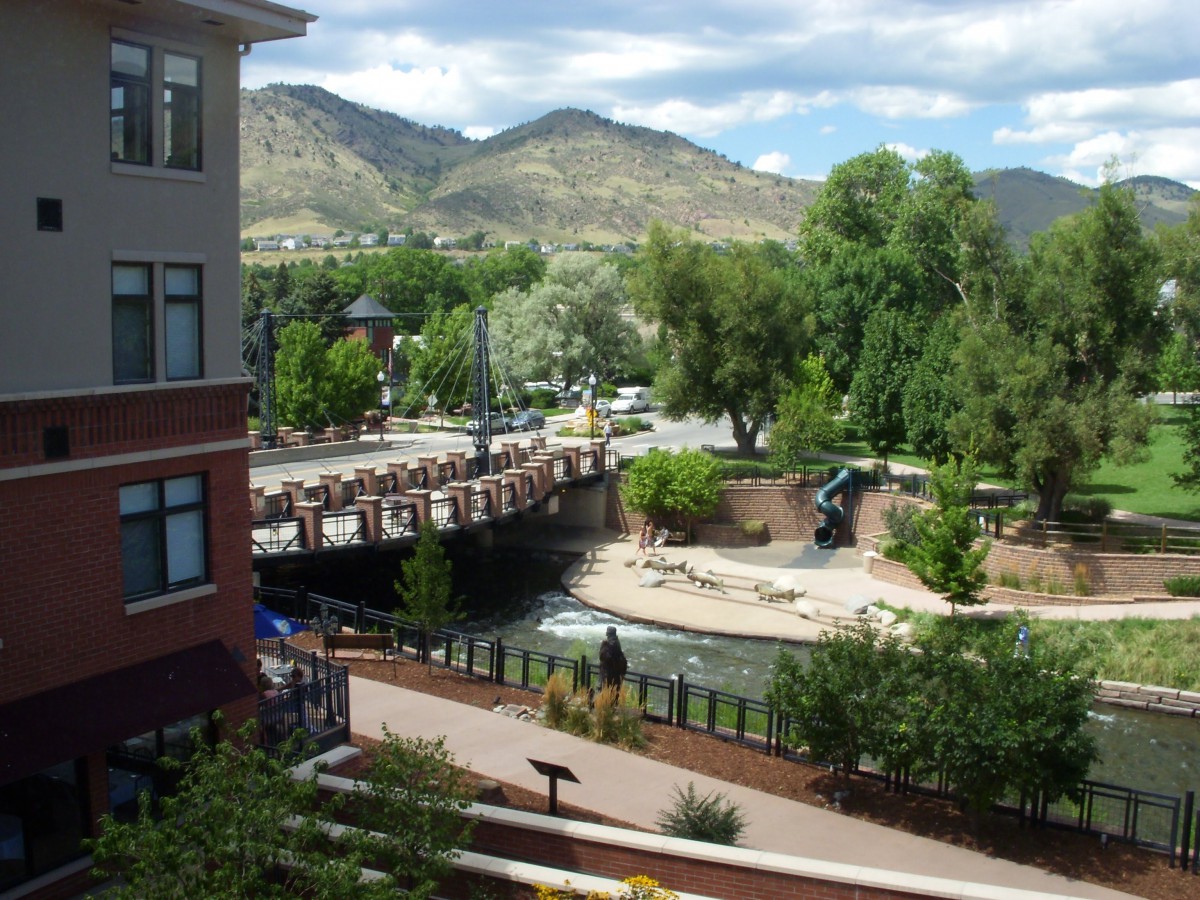 A scenic view of a river flowing under a bridge with mountains in the background, surrounded by trees and buildings.